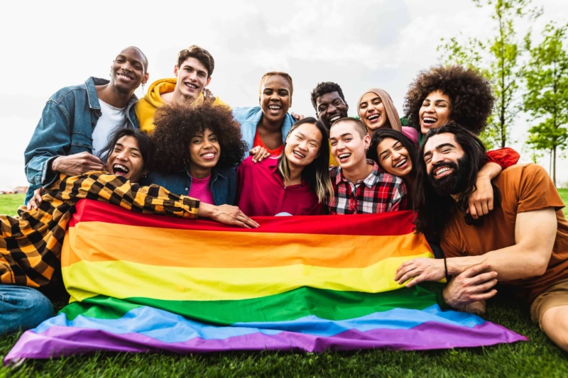 A group of happy people, holding a pride flag while smiling. The flag is the 6-color pride flag.